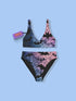 Model wearing a high-waisted pastel goth bikini set featuring a black bat and spiderweb print over an ombré pink and purple background. The two-piece swimsuit includes a scoop neck top and high-rise bottoms, styled outdoors in bright festival and poolside settings. Made of recycled fabric, soft, stretchy, and UV resistant. Available in XS to 3X. Aesthetic is gothic, spooky cute, and Y2K inspired.